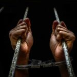 Hands in handcuffs gripping jail bars.
