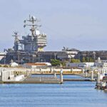 A large naval aircraft carrier docked in a harbor with smaller boats in the foreground