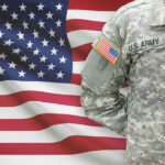 Soldier in camouflage uniform standing before American flag.