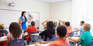 Teacher in a blue dress instructing students in a classroom with hands raised