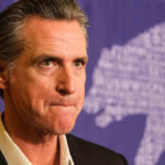Newsom’s Top Aide ARRESTED – Faces 23 Counts! Man in suit, solemn expression, purple background with bear drawing.