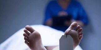 A deceased body on a table with a tag on the foot, and a medical professional in the background