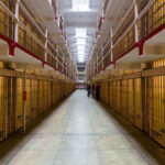 Torture Uprising – Prison Inmates Ignite Matresses! Long hallway with prison cells on both sides.