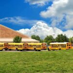 A row of yellow school buses parked in front of a school building under a cloudy sky