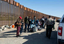 Group of people standing by a border fence with officers and vehicles