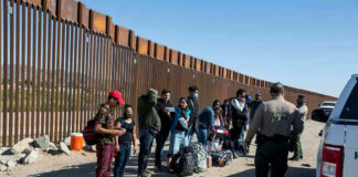 Group of people standing by a border fence with officers and vehicles