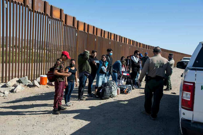 1973414822 Group of people standing by a border fence with officers and vehicles