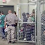 Detention facility with people behind chain-link fence and officer nearby
