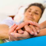 Biden Cancer Spreads – Family Prepares For The Worst Caregiver holding hands with an elderly patient in bed