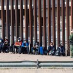Group of people sitting by a tall border fence with an officer standing nearby