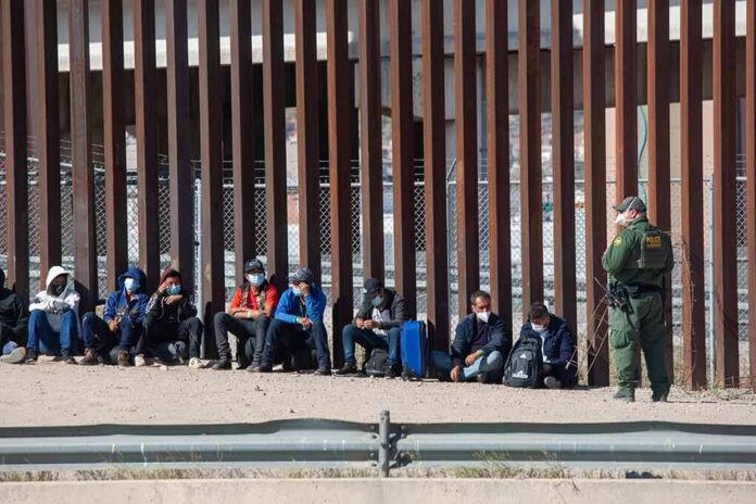 shutterstock_2079408472-1 Group of people sitting by a tall border fence with an officer standing nearby