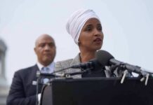 MTG Comes To Arch Nemesis’s DEFENCE Ilhan Omar speaking at a podium with André Carson standing behind