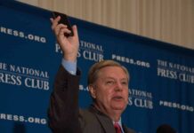 Man in suit speaking at podium holding up a smartphone